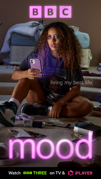 A woman with curly hair sits indoors holding a phone, posing in front of a bed and floor covered with objects, for a 'Mood' TV show ad.