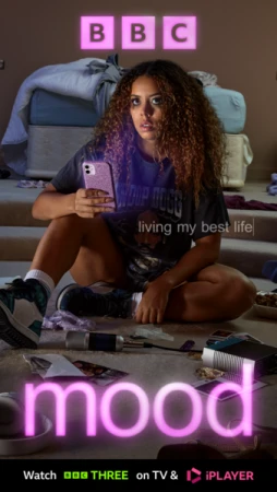A woman with curly hair sits indoors holding a phone, posing in front of a bed and floor covered with objects, for a 'Mood' TV show ad.