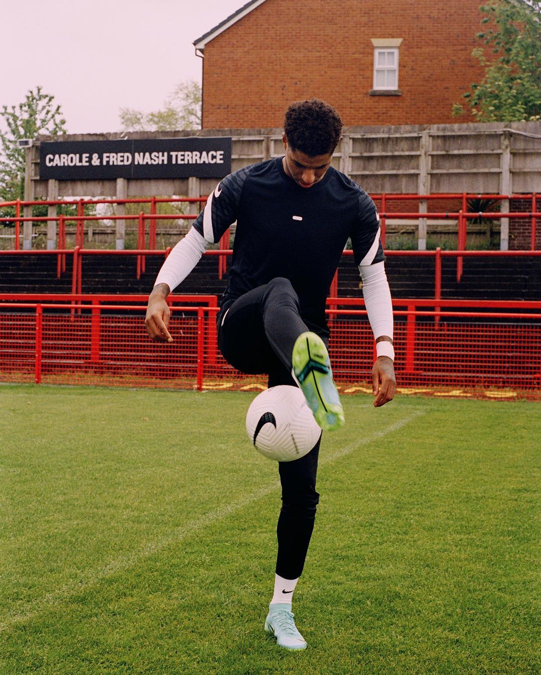 Adult male athlete in sportswear balances a soccer ball on his foot on a green field in front of red bleachers and a brick building.