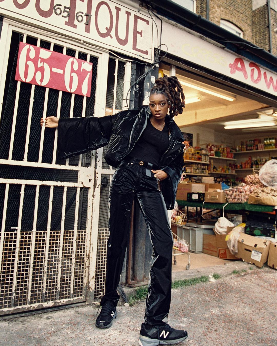 A young woman poses in black puffer jacket and pants outside a shop with a grey exterior. She wears New Balance shoes.