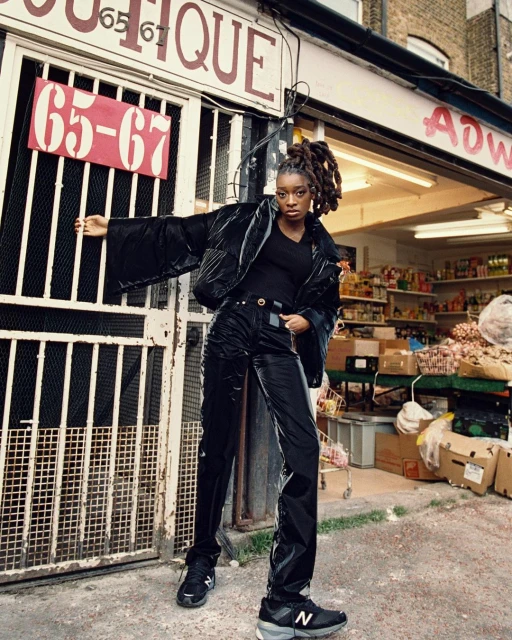 A young woman poses in black puffer jacket and pants outside a shop with a grey exterior. She wears New Balance shoes.