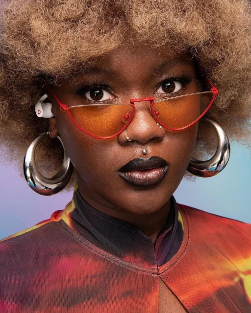 Portrait of a stylish young Black woman wearing orange geometric sunglasses and large silver hoop earrings against a colorful gradient backdrop.