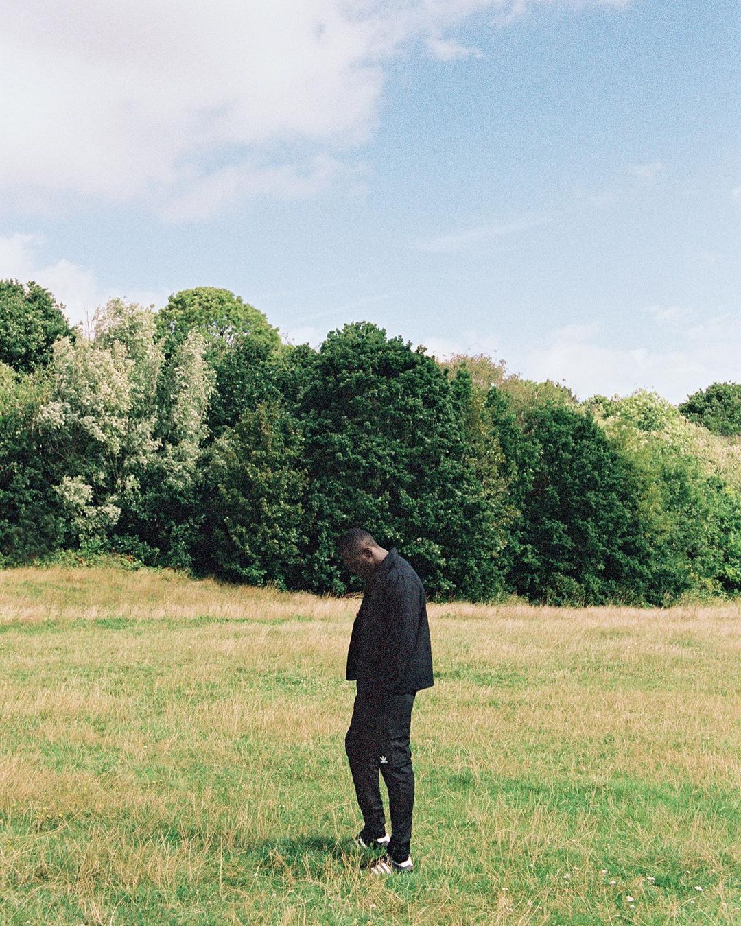 A dark-skinned man stands in a grassy field with trees and a cloudy sky in the background. He is wearing a dark tracksuit.