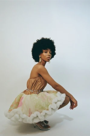 Model with afro hairstyle crouching in strapless dress against a white background