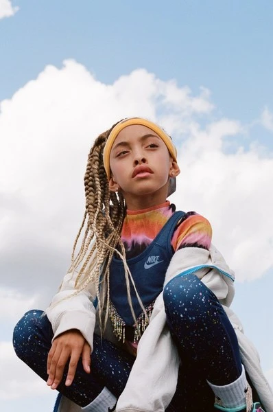 Portrait of a young girl wearing a colorful t-shirt, blue overalls, and a yellow headband against a cloudy sky backdrop. The girl is squatting.