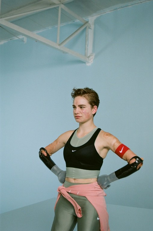Athlete in sportswear poses in front of light blue wall. She wears arm supports, crop top, leggings, and pink sweater.