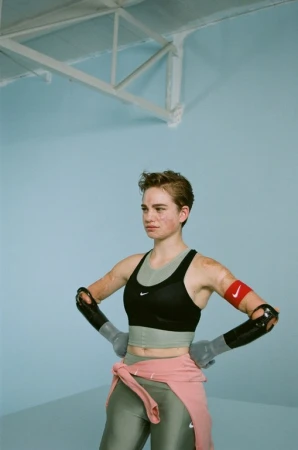 Athlete in sportswear poses in front of light blue wall. She wears arm supports, crop top, leggings, and pink sweater.