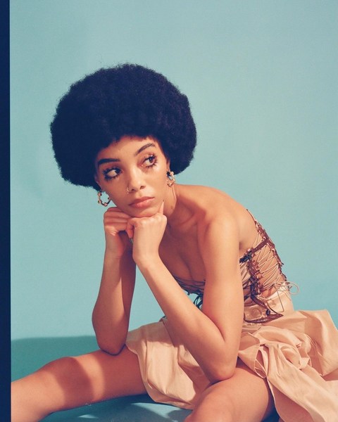 Portrait of an adult woman with an afro hairstyle sitting in a photo studio wearing a strapless top and skirt against an aqua background.