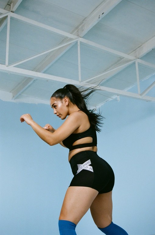 Athletic woman in boxing stance in gym. She wears black athletic shorts and top with blue knee-high socks on blue background.