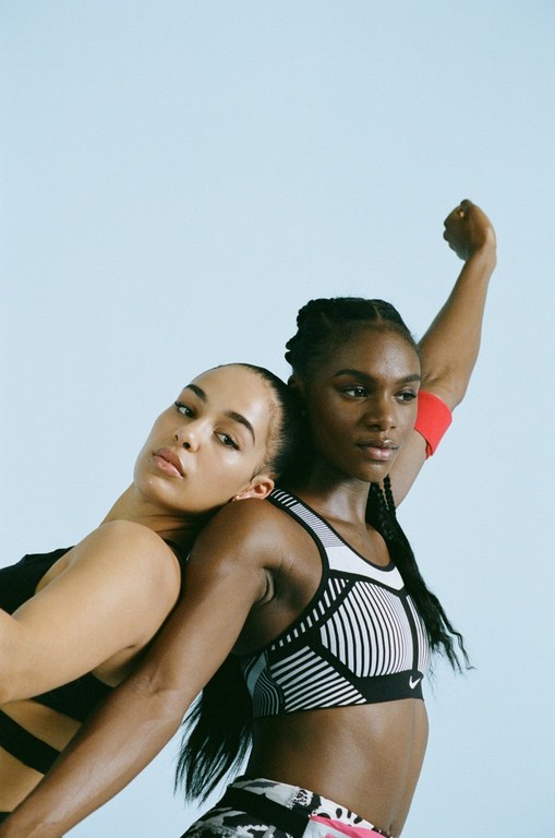 Two athletic women posing back-to-back wearing sports bras against a light blue background. One has her arm raised.