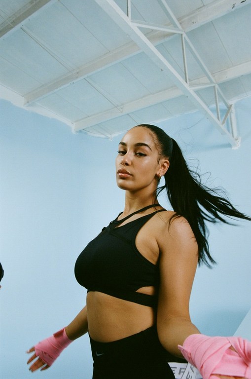 Athletic woman in sports bra with pink hand wraps prepares for workout in a gym setting with a modern, minimalist aesthetic.
