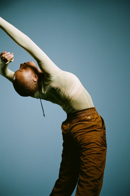 Full-length portrait of a person with arms raised and arched back, wearing a sheer long-sleeve shirt and brown pants, against a blue background.