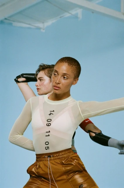 Two athletic women with prosthetic arms posing indoors. One wears an off-white long-sleeve shirt, copper shorts, and Nike logo visible.