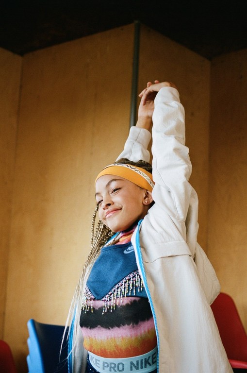 Smiling young girl in a colorful sportswear outfit stretches her arms against a yellow and brown wall indoors. Braids and headband visible.
