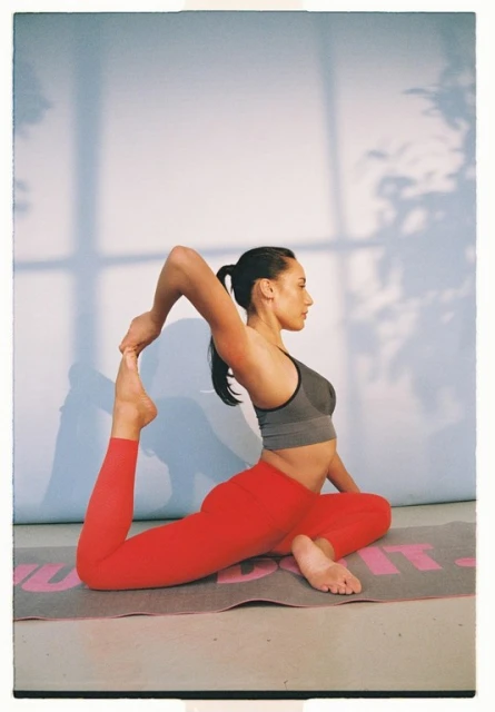 A woman in a grey sports bra and red leggings is performing a yoga pose while holding her foot on a mat in an indoor setting.