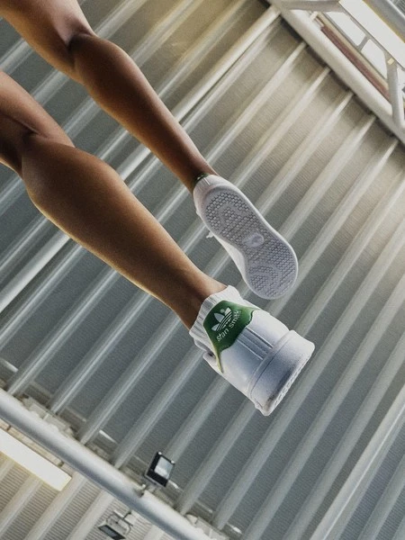 Low-angle shot showcasing legs and feet of a person wearing white Adidas Stan Smith sneakers on a metal ceiling background.