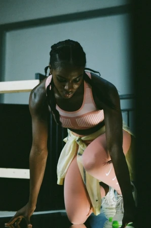 Focused female athlete in sportswear kneeling, poised for action. Fitness photoshoot in indoor athletic setting with determination.