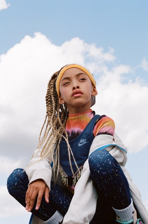 A young person wearing a colorful outfit and yellow headband squats against a bright sky, shot from a low angle.