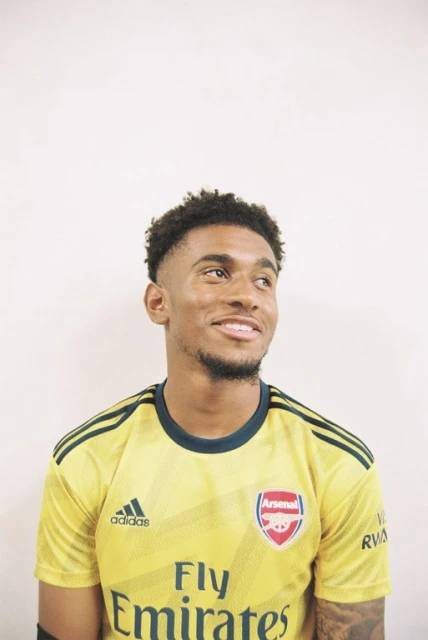 Smiling man in a yellow Arsenal soccer jersey stands against a white backdrop, looking off to the right side of the frame.