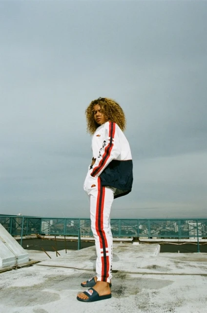 Woman with curly hair, wearing a white and red tracksuit with sandals, stands on a rooftop with a hazy sky, looking over her shoulder.