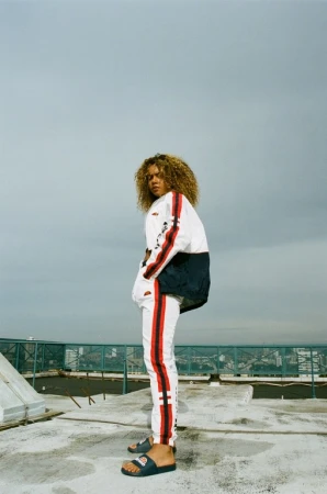 Woman with curly hair, wearing a white and red tracksuit with sandals, stands on a rooftop with a hazy sky, looking over her shoulder.