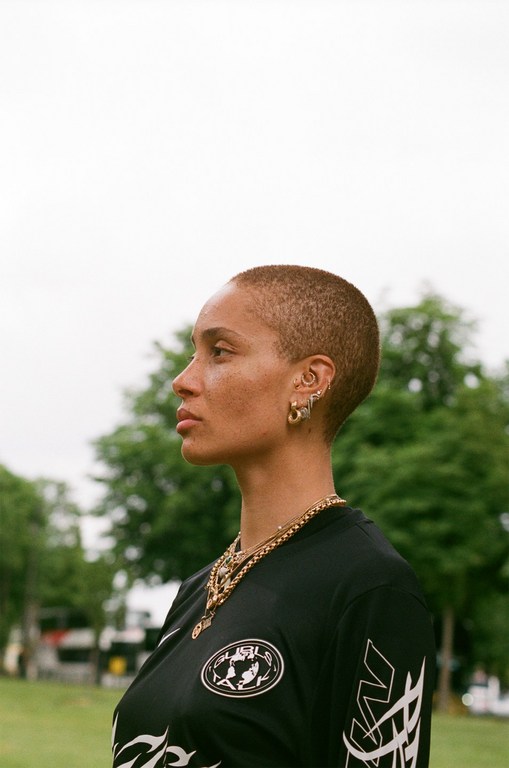 Profile shot of a woman with short hair and gold jewelry outdoors. She wears a black soccer jersey with white and gold graphic accents.