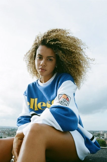 A woman with curly hair poses on a rooftop wearing a vintage Ellessee sweatshirt against a cloudy sky and cityscape backdrop.