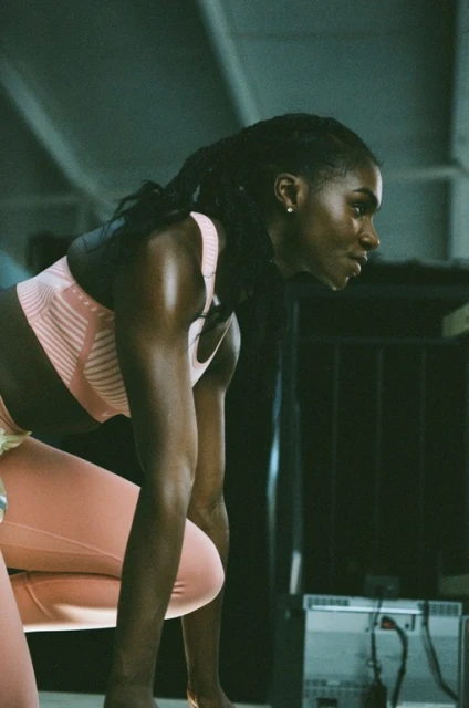 Athletic Black woman in a pink sports bra and leggings crouches in an indoor workout space with overhead lighting and an exercise machine.