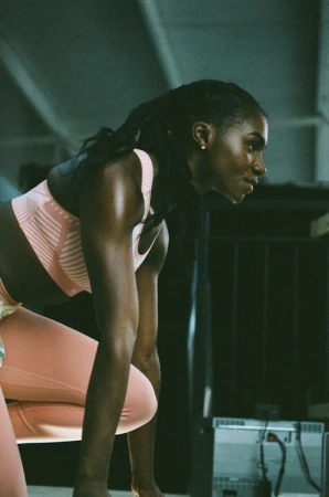 Athletic Black woman in a pink sports bra and leggings crouches in an indoor workout space with overhead lighting and an exercise machine.