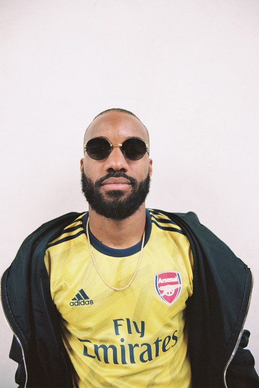 Bearded man in round sunglasses wearing a yellow soccer jersey with a jacket against a white background. Fashion portrait.