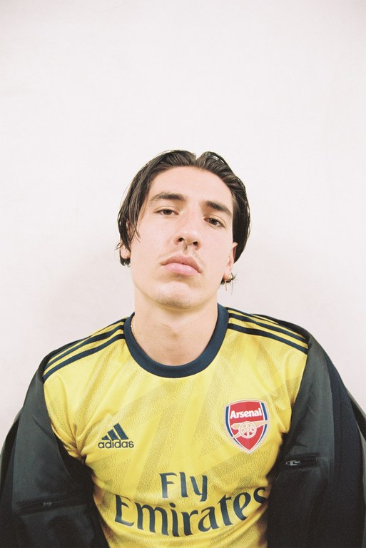 A young adult male poses indoors, wearing a yellow and blue Arsenal soccer jersey with visible logos, in a medium shot with a white backdrop.
