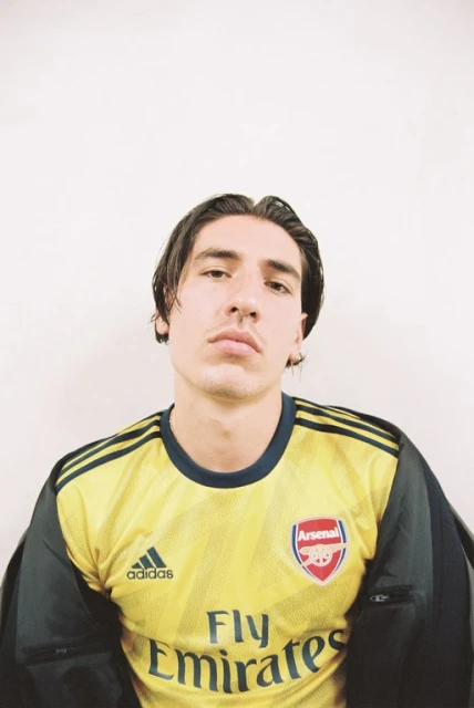 A young adult male poses indoors, wearing a yellow and blue Arsenal soccer jersey with visible logos, in a medium shot with a white backdrop.