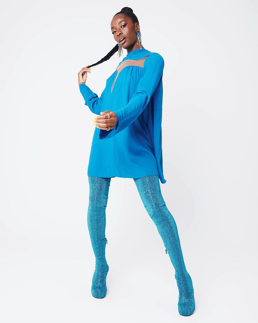 Fashion shot of an African woman wearing a blue dress and sparkly blue tights in a white studio setting, posing and holding her hair.