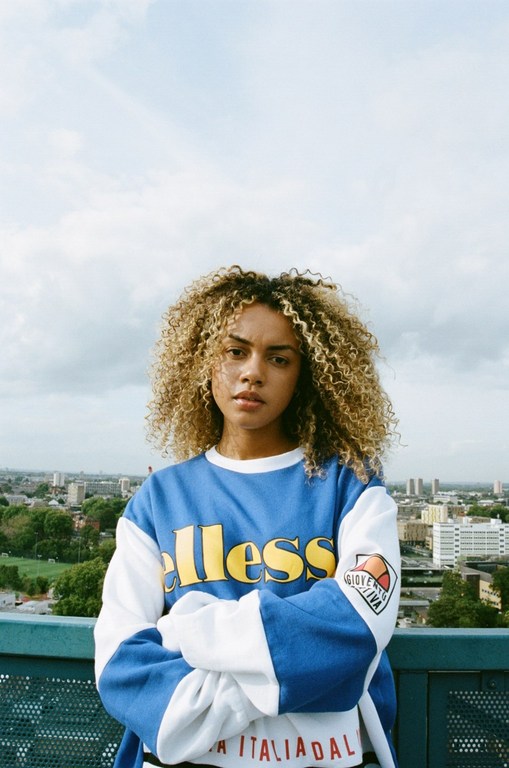 Woman with curly blonde-highlighted hair wearing a blue and white Ellesse sweatshirt with arms crossed against a cityscape rooftop background.