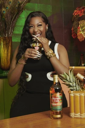 Smiling African American woman in a black-and-white dress drinking a mixed drink next to a bottle of Cuban Spiced Havana Club rum.