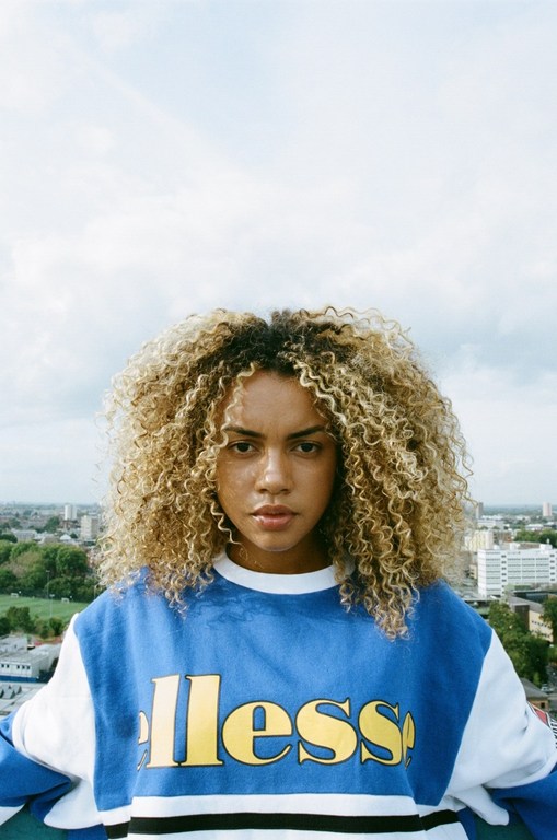 Portrait of an adult woman with curly hair, wearing a blue and white sweatshirt with a logo, against an urban and cloudy sky backdrop.