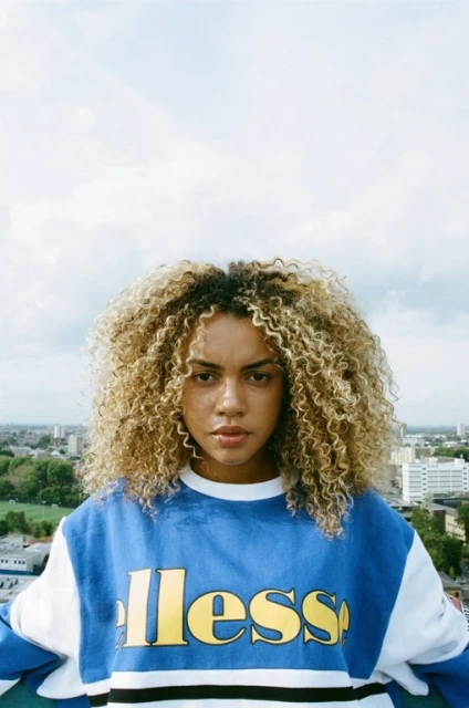 Portrait of an adult woman with curly hair, wearing a blue and white sweatshirt with a logo, against an urban and cloudy sky backdrop.