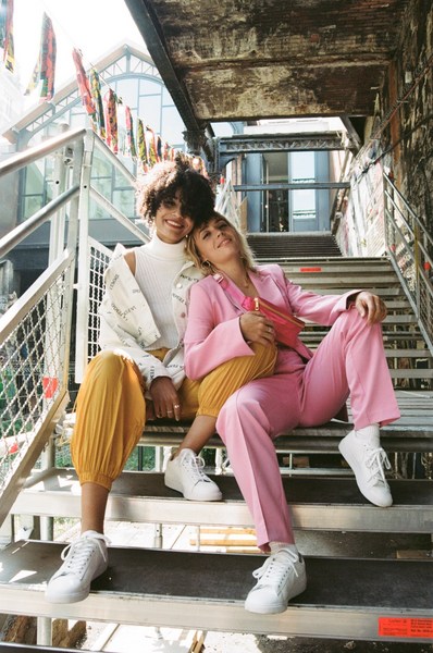 Two women in fashionable outfits sit on outdoor steps, one in a pink suit, the other in yellow pants and a white jacket, smiling at the camera.