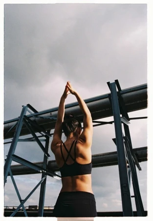 Back view of a sporty woman stretching arms upwards on a rooftop with industrial pipes and a cloudy sky background in an outdoor setting.