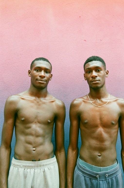 Two shirtless young Black men posing together against a textured pink and blue gradient wall in London by Danika Magdelena