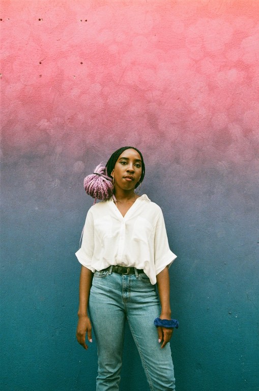 Woman poses in front of an ombre-painted wall, wearing a white button-down shirt, jeans, and a lavender wrap on her head.