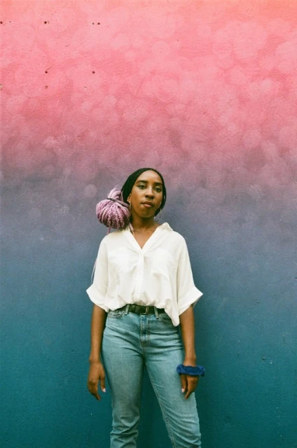 Woman poses in front of an ombre-painted wall, wearing a white button-down shirt, jeans, and a lavender wrap on her head.