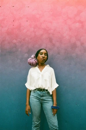 Woman poses in front of an ombre-painted wall, wearing a white button-down shirt, jeans, and a lavender wrap on her head.