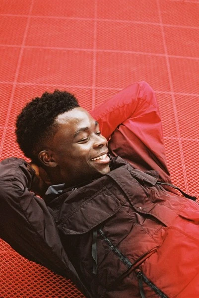 Smiling young man with short black hair lies on red geometric surface wearing a dark jacket; low angle, medium shot with soft lighting.
