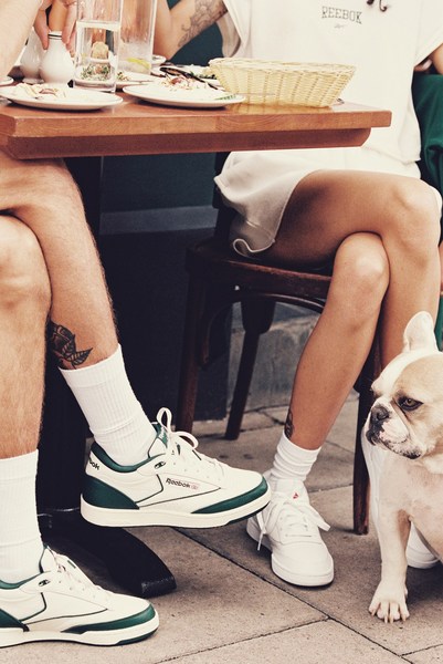 Close-up of a man and woman seated at a wooden table, both wearing white Reebok sneakers, with a French Bulldog dog visible.