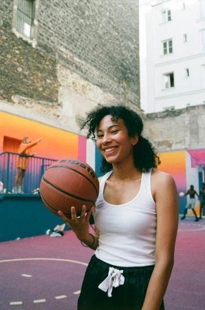 Smiling woman in a white tank top holding a basketball on a colorful outdoor basketball court in London by Danika Magdelena