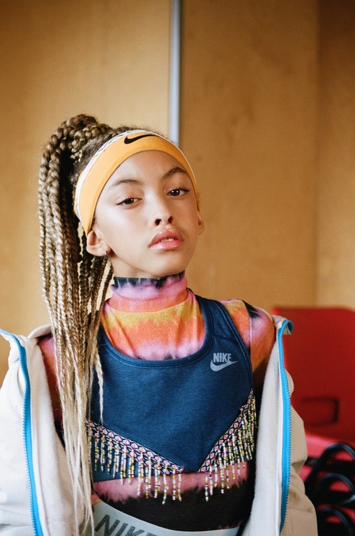 Portrait of a young girl with blonde braids wearing a colorful t-shirt, sports bra, and a Nike headband in front of a tan wall.