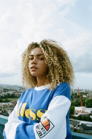 Woman with curly blonde hair wearing a blue and white sweatshirt posing outdoors against a skyline