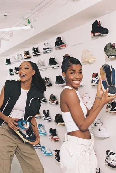 Two smiling women showcase sneakers in a retail setting with a wall display of various shoes, presenting a curated collection.