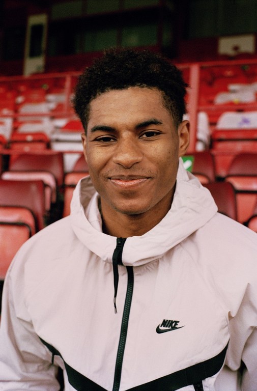 Close-up shot of a smiling, dark-skinned man in a light pink Nike hooded jacket, sitting in a red stadium seat. Sport and fashion.